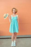 young cheerful curly redhead woman in blue sundress holding straw hat in her hand on beige background. Fun, summer, fashion, youth concept