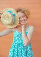 young cheerful curly redhead woman in blue sundress holding straw hat in her hand on beige background. Fun, summer, fashion, youth concept photo