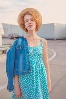 joven pelirroja rizada con sombrero de paja, vestido azul y chaqueta de jeans de pie en el techo moderno. diversión, verano, moda, tiro, viajes, concepto juvenil. espacio de copia foto