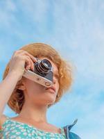 young curly redhead woman in straw hat, blue sundress and jeans jacket standing with vintage camera and taking pictures on blue sky background. Fun, summer, fashion, shooting, travel, youth concept photo