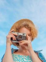 young curly redhead woman in straw hat, blue sundress and jeans jacket standing with vintage camera and taking pictures on blue sky background. Fun, summer, fashion, shooting, travel, youth concept