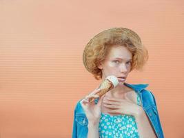 young curly redhead woman in straw hat, blue sundress and jeans jacket eating ice cream on beige background. Fun, summer, fashion, youth concept photo