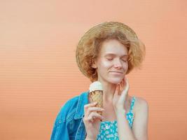 young curly redhead woman in straw hat, blue sundress and jeans jacket eating ice cream on beige background. Fun, summer, fashion, youth concept photo