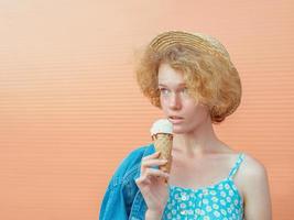 young curly redhead woman in straw hat, blue sundress and jeans jacket eating ice cream on beige background. Fun, summer, fashion, youth concept photo