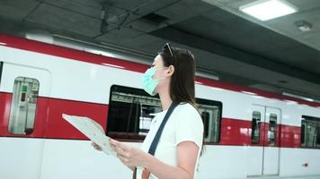 Beautiful Asian female tourist with face mask, eyeglasses, and camera,  searching traveler location map, looking for destination at train station in Thailand, railway transportation, casual lifestyle.