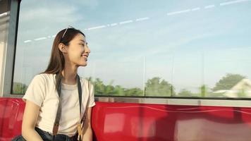 una hermosa turista asiática con cámara se sienta en un asiento rojo, viaja en tren, toma una foto instantánea cuando se transporta a la vista urbana, disfruta del estilo de vida de los pasajeros en tren, feliz viaje de vacaciones. video