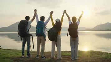 cuatro hombres y mujeres adultos asiáticos que llevan un paquete de viaje, viajan hasta el final, disfrutan de su día y se relajan con las manos en alto. durante la puesta de sol. tiene vista al embalse y montañas al fondo. video