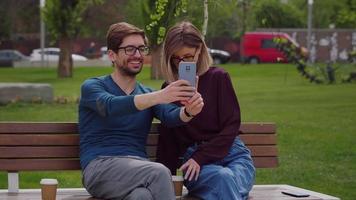 Two friends sit on the bench outside take selfie with phone. video