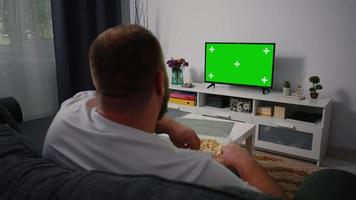 achteraanzicht man zittend op de bank in de woonkamer tv kijken met groen scherm en kanalen veranderen met afstandsbediening in zijn hand. video