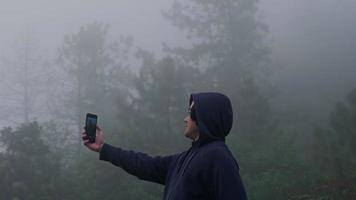 ung man resenär i marinblå manlig huvtröja lång ärm med hjälp av en smartphone och njut av landskapet i bergen med tjock dimma i bakgrunden. video