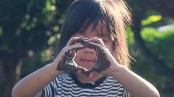 Cute little girl with muddy hands makes a heart shape with her hands against nature background. Concept of nature and love symbol. video