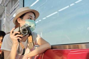 Beautiful Asian female tourist with face mask sits in a red seat, traveling by train, taking snapshot photo, transporting in suburb view, enjoy passenger lifestyle by railway, happy journey vacation. photo