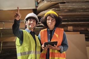 Two female workers and colleagues in safety uniforms and helmet stock check, control production in storage of warehouse factory with a lot of paper stack, friend works in recycle manufacture business. photo