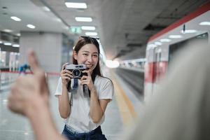 joven y hermosa turista asiática tomando una foto de retrato con una cámara de cine con un amigo, sonriendo y disfrutando en la plataforma de la estación de tren, feliz estilo de vida de viaje en viaje de vacaciones en transporte subterráneo.