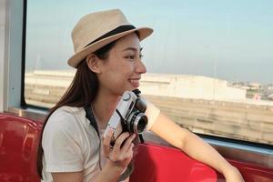 Beautiful Asian female tourist sits in a red seat, traveling by train, taking snapshot photo, transporting in suburb view, enjoy passenger lifestyle by railway, happy journey vacation. photo