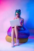 a woman in a bright bathing suit with blue hair is sitting on a chair bag with a laptop photo