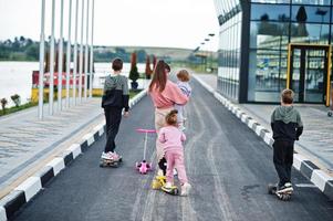 Back of young stylish mother with four kids outdoor. Sports family spend free time outdoors with scooters and skates. photo