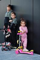 Three kids outdoor against black modern wall. Sports family spend free time outdoors with scooters and skates. photo