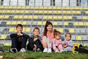 joven madre elegante con cuatro hijos sentados en el césped contra el estadio. la familia deportiva pasa tiempo libre al aire libre con scooters y patines. foto