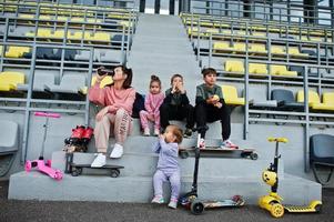 joven madre elegante con cuatro hijos sentados en el podio deportivo en el estadio, come manzana y bebe agua. la familia pasa tiempo libre al aire libre con scooters y patines. foto