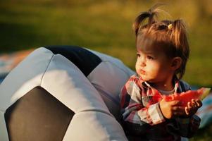 Cute baby girl in checkered shirt sit on football ball pouf and eat watermelon. photo