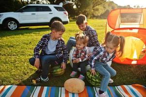 Four kids spending time together. Outdoor picnic blanket, sitting with watermelons. photo