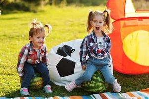 Two little cutie sister girls outdoor in picnic, wear checkered shirt. Sitting on watermelon. photo