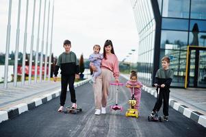 Young stylish mother with four kids outdoor. Sports family spend free time outdoors with scooters and skates. photo