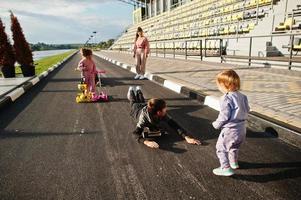 Young stylish mother with four kids outdoor. Sports family spend free time outdoors with scooters and skates. photo