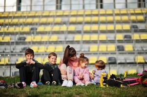 joven madre elegante con cuatro hijos sentados en el césped contra el estadio. la familia deportiva pasa tiempo libre al aire libre con scooters y patines. foto