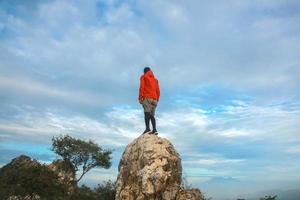 Man hiker with orange jacket standing on top of stone and enjoying amazing view on bright blue sky. Travel and active lifestyle concept. photo
