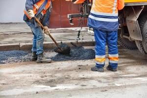 Road workers with shovels stand next to a truck pouring fresh asphalt onto a repaired section of road. photo
