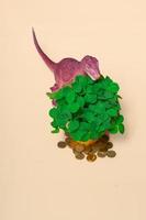 St. Patrick's Day happy dinosaur, a bouquet of clover in a bucket and gold coins on a beige background. Vertical format, closeup copy space photo