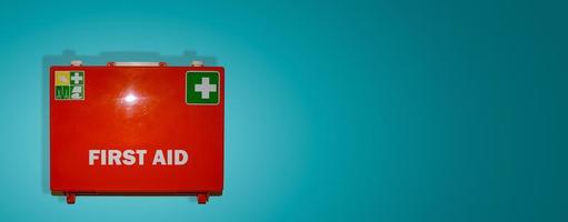 Banner with orange first aid case labeled in English language First Aid hanging on the wall for safety measures, at turquoise gradient background copy space. Concept of emergency medical assistance. photo