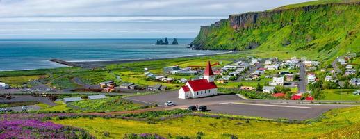 Panoramic view of basalt stacks Reynisdrangar, volcanic black sand beach and violet lupine and yellow meadow flowers at Vik town, and a Lutheran church, South Iceland, at summer sunny day blue sky.