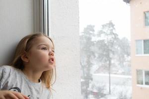A little girl looks out the window at the flying snow and the winter snow-covered town photo