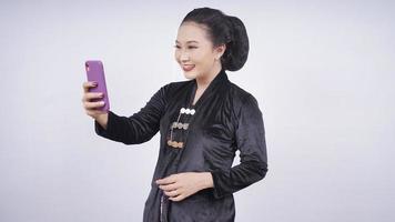 Belleza asiática en kebaya jugando con celular aislado sobre fondo blanco.