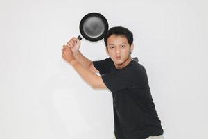 Young Asian man is angry and ready to strike with a frying pan. Indonesia Man wear black shirt Isolated grey background photo