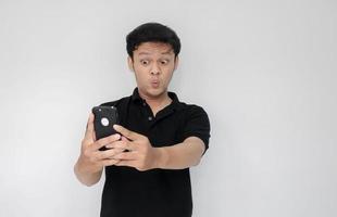 wow, la cara de tu hombre asiático se sorprendió por lo que vio en el teléfono inteligente con un fondo gris aislado. indonesia hombre usar camisa negra aislado fondo gris foto