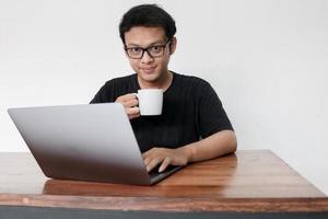 Young asian man is smile and enjoy when working at home with laptop and a cup of coffee. A man sitting on bed and working. Work from home concept photo