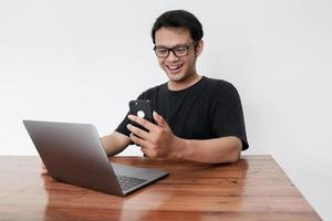 Young Asian man feeling happy and smile what he see in the smartphone and laptop on the table. Indonesia Man wear black shirt Isolated grey background. photo