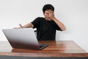 Young asian man hiding his face with hand because shocked and embarrassed by some porn videos or another forbidden thing he saw on the internet using a laptop photo