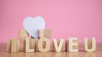 These words mean I LOVE U in wood plates. The square blocks support the white heart. Close up shot isolated on pink background. valentine's day concept. photo