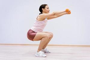 Woman squats with dumbbells in her hand against the background of a light wall photo