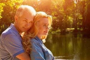 feliz pareja madura de pie en un abrazo en el lago bajo el sol foto