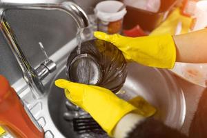 Close up of female hands in yellow protective rubber gloves washing white plate with blue cleaning sponge against kitchen sink full of foam and tableware