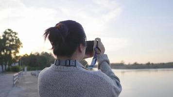 bakifrån av asiatisk kvinna som står och tar en bild vid floden i vacker dag. håller i kameran och tar ett foto. floden bakgrund. avkopplande dag på kvällen video