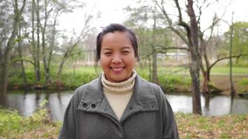 close up front view of Asian woman standing at the forest, smiling and looking at camera. Autumn season, leaves falling from the trees , beautiful nature, river background, Sweden video