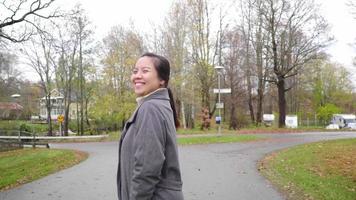 glückliche asiatische frau im park, lächelnd und blick in die kamera. beim Gehen umdrehen. herbstsaison, blätter fallen von den bäumen, schöne natur, frische luft im park, schweden video