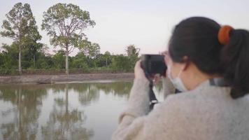 bakifrån närbild av asiatisk kvinna som står och tar en bild av båten vid floden på kvällen. håller kamera med floden bakgrund. avkopplande dag video